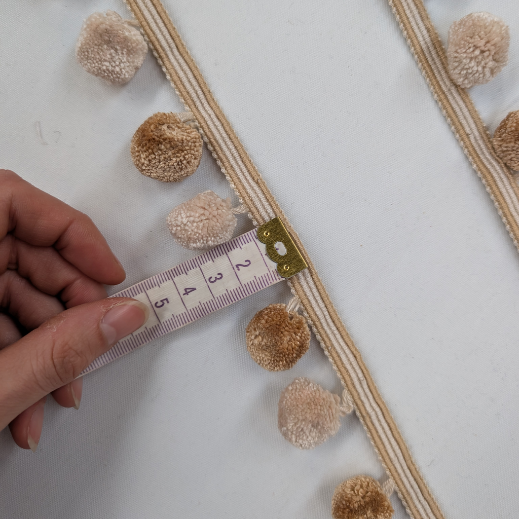 Hand measuring a decorative ribbon with pom-poms using a tape measure on a light background