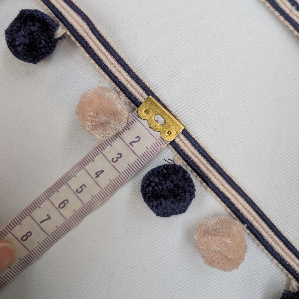 Close-up of a decorative ribbon with pom-poms and a gold clasp, measured against a ruler.